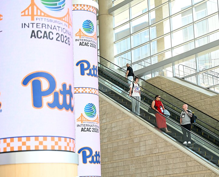 People ride an escalator next to columns with Pitt and International ACAC logos.