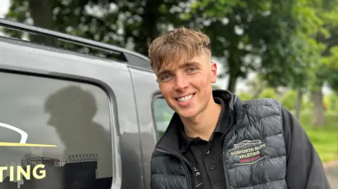 BBC Louis Clougher has brown hair which is longer on top and cropped to the sides and wears a black padded gilet with his company's branding. He is leaning against one of his valet vans and smiling.