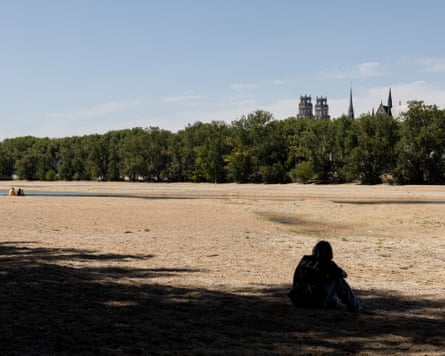 People sit in the dry, brown Loire riverbed with only a tiny trickle of water seen. The spires and turrets of Orleans are in the background behind a line of trees on the opposite riverbank.