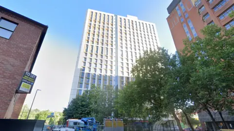 Google A city street lined with tall buildings, including a modern white‑and‑glass high‑rise, an older brick building with a large “To Let” office‑space sign, and a reddish‑brown tower. Trees partially obscure the view, with construction vehicles and equipment at street level.
