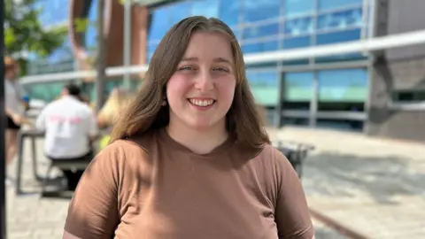 Florence McMahon has long brown hair and is wearing a brown coloured t-shirt. She is pictured smiling outside an office building.