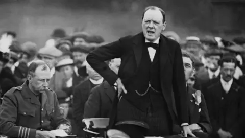 Hulton-Deutsch Collection/CORBIS/Corbis via Getty Images Sir Winston Churchill, wearing a tuxedo, addresses a crowd from a raised stage. He has one hand on his hip.