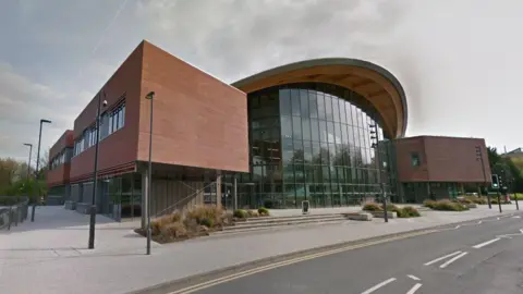 Google A large, glass-front building at the University of Warwick. The pavement and road in front of the building are also visible, and bushes line the steps and entrances to the building.