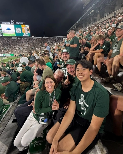 Logan Rizal, foreground right, smiles with fellow fans in the crowded stands at Spartan Stadium during a nighttime football game.
