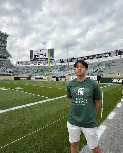 Logan Rizal stands beside the football field at Spartan Stadium wearing a green “Global Spartan” T-shirt and white shorts.