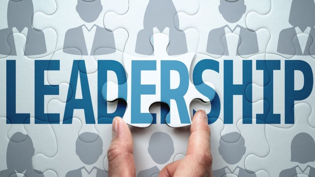 Hands lift a puzzle piece to reveal the word LEADERSHIP, with silhouettes of people in the background.