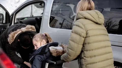 Getty Images A person with long blonde hair, wearing a khaki green puffer jacket, with her back to the camera, appears to hold a wheelchair frame with a woman or girl sitting on it. Behind her is a large silver transit vehicle.