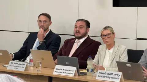 Simon Dedman/BBC Sam Journet sits behind a table, with a laptop open in front of him. He wears a velvet-looking red suit and pink tie. He is flanked by a man on his right, and a woman with blonde hair to his left.