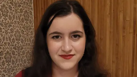 Lancashire Police Carla looks at the camera. She has brown eyes and thick long dark hair. She is wearing red liptstick and there is a wooden wall behind her.