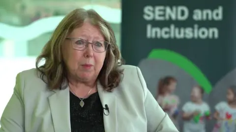 Councillor Elaine Lynch. She has shoulder-length blond hair and blue eyes. She wears glasses with a thin metal rim. She is wearing a cream blazer and sparkly black top and a silver pendant around her neck. She is looking at the camera with a serious expression. In the blurred background there is a banner with the words "SEND and inclusion"