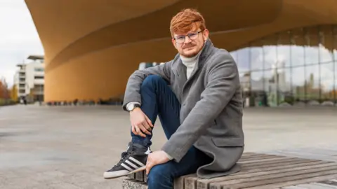 Owein Iveson Owein Iveson sits outside, wearing a grey coat, blue jeans and trainers